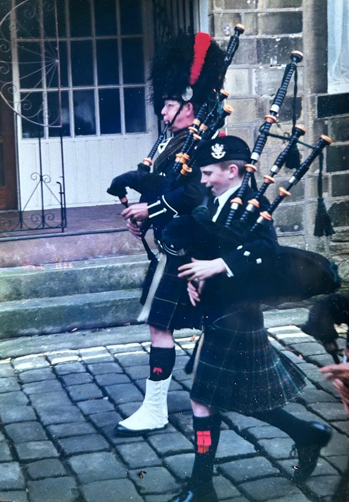 Young James Gore playing bagpipes alongside an adult piper in Highland dress